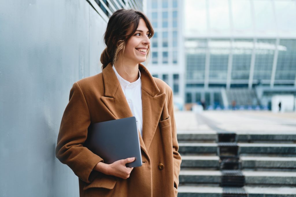Young cheerful businesswoman in coat with laptop happily looking away on city street